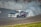 A sports car is executing a high-speed drift around a racetrack, creating a large cloud of white smoke from its tires. The car is adorned with various decals and sponsors, and spectators can be seen watching from behind a fence in the background.