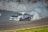 A sports car is executing a high-speed drift around a racetrack, creating a large cloud of white smoke from its tires. The car is adorned with various decals and sponsors, and spectators can be seen watching from behind a fence in the background.