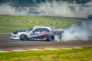 A racing car performing a drift on a track with smoke billowing from the rear tires. The car displays vibrant graphics, including the words 'MIDNIGHT' along the side. The background features a crowd of spectators blurred by the smoke.