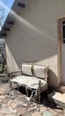 An old, damaged couch with torn and worn upholstery is placed outside against the wall of a building. Sunlight casts shadows, highlighting the decay of the furniture. The ground is covered with broken stones and debris, and there is a small patch of grass nearby. A door with a barred window is partially visible, framed by the building's rough, beige exterior.