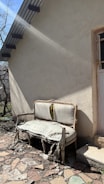 An old, damaged couch with torn and worn upholstery is placed outside against the wall of a building. Sunlight casts shadows, highlighting the decay of the furniture. The ground is covered with broken stones and debris, and there is a small patch of grass nearby. A door with a barred window is partially visible, framed by the building's rough, beige exterior.