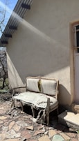 An old, damaged couch with torn and worn upholstery is placed outside against the wall of a building. Sunlight casts shadows, highlighting the decay of the furniture. The ground is covered with broken stones and debris, and there is a small patch of grass nearby. A door with a barred window is partially visible, framed by the building's rough, beige exterior.