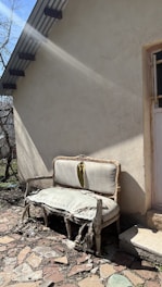 An old, damaged couch with torn and worn upholstery is placed outside against the wall of a building. Sunlight casts shadows, highlighting the decay of the furniture. The ground is covered with broken stones and debris, and there is a small patch of grass nearby. A door with a barred window is partially visible, framed by the building's rough, beige exterior.