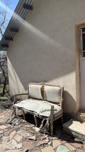 An old, damaged couch with torn and worn upholstery is placed outside against the wall of a building. Sunlight casts shadows, highlighting the decay of the furniture. The ground is covered with broken stones and debris, and there is a small patch of grass nearby. A door with a barred window is partially visible, framed by the building's rough, beige exterior.