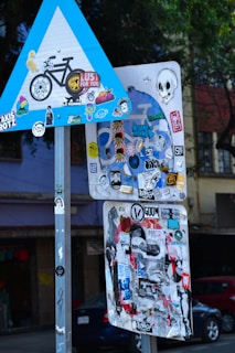 A collection of road signs covered with various colorful stickers, including designs of skulls, faces, and patterns, is seen against an urban backdrop with trees and buildings.