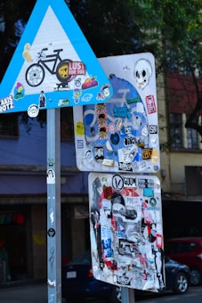 A collection of road signs covered with various colorful stickers, including designs of skulls, faces, and patterns, is seen against an urban backdrop with trees and buildings.