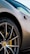 A close-up view of a luxury sports car wheel, featuring a Pirelli tire and a sleek, multi-spoke rim with a yellow brake caliper. The car’s body is elegantly curved and has a metallic finish.