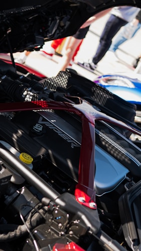 Close-up of a car engine bay with a visible Viper logo on the engine cover. The components are encased by a red strut brace. A blue car is partially visible in the background, and there are people standing around in casual clothing.