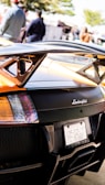 The image features the rear section of a black Lamborghini sports car, focusing on its distinctive taillights and rear wing. The license plate is visible, and the Lamborghini logo is prominently displayed. In the background, a few people are gathered, slightly blurred, indicating the car might be showcased at an outdoor event.