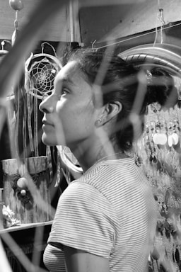 A black and white photograph of a person in profile, surrounded by dreamcatchers and artistic decorative elements. The focus is on their contemplative expression, while the background includes intricate designs and textures.