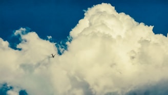 A large, fluffy white cloud dominates the sky with a deep blue backdrop. In the center-left, a small airplane is visible, contrasting against the bright cloud.
