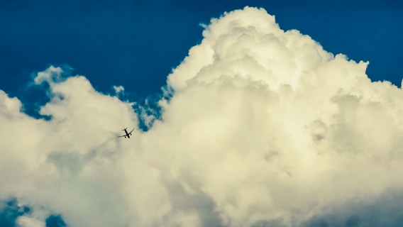 A large, fluffy white cloud dominates the sky with a deep blue backdrop. In the center-left, a small airplane is visible, contrasting against the bright cloud.