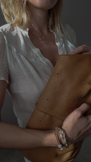 A person with blond hair is holding a brown, textured leather handbag. They are wearing a white, short-sleeved top with subtle patterns. A delicate necklace with a circular pendant is visible around their neck. On the wrist, there is a bracelet featuring a chain and some beads.
