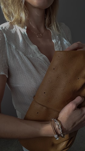 A stylish young woman wearing layered gold necklaces and holding a nude-toned handcrafted bag.