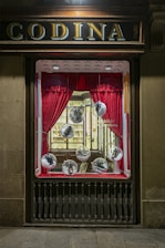 A vintage storefront display features red curtains framing large glass spheres, each containing a shoe. The backdrop includes a well-lit, elegant interior with wooden shelves and a display of various items, creating a classic and sophisticated atmosphere.