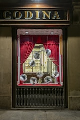 A vintage storefront display features red curtains framing large glass spheres, each containing a shoe. The backdrop includes a well-lit, elegant interior with wooden shelves and a display of various items, creating a classic and sophisticated atmosphere.