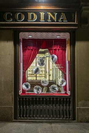 A vintage storefront display features red curtains framing large glass spheres, each containing a shoe. The backdrop includes a well-lit, elegant interior with wooden shelves and a display of various items, creating a classic and sophisticated atmosphere.