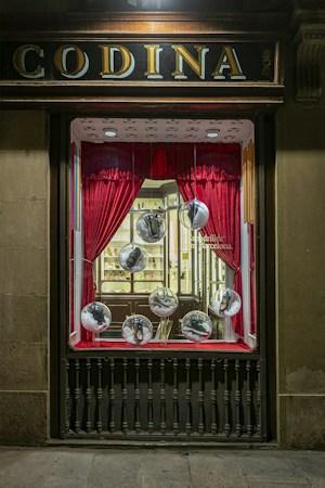 A vintage storefront display features red curtains framing large glass spheres, each containing a shoe. The backdrop includes a well-lit, elegant interior with wooden shelves and a display of various items, creating a classic and sophisticated atmosphere.