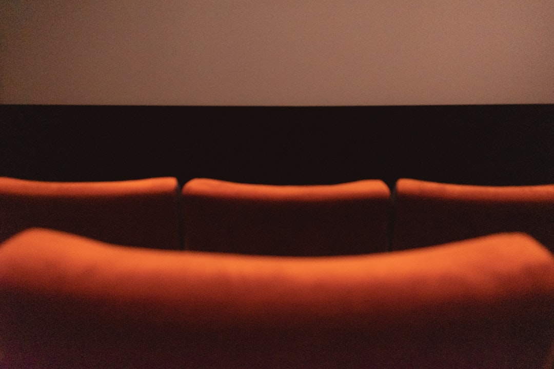 a row of orange couches in front of a projector screen, Back view of red seats in a theater.