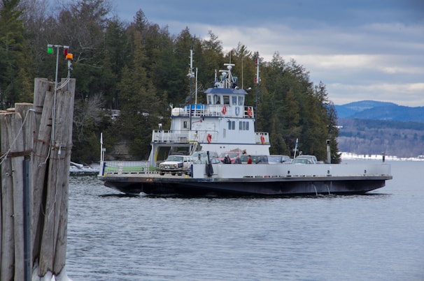 A large ferry boat is transporting several cars across a body of water. The scene is set against a backdrop of dense, evergreen forest lining the shore. The sky above is overcast, casting a greyish hue over the landscape. Some mountains can be seen in the distance, adding depth to the scene.