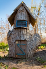 A whimsical tree trunk structure resembling a small house, complete with a wooden door and a window framed in blue. The roof is made of wooden planks, giving it a rustic appearance. It is set outdoors in a natural setting with green foliage and trees in the background.