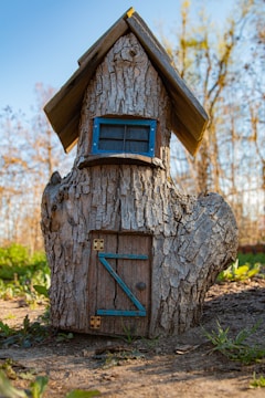 A whimsical tree trunk structure resembling a small house, complete with a wooden door and a window framed in blue. The roof is made of wooden planks, giving it a rustic appearance. It is set outdoors in a natural setting with green foliage and trees in the background.