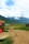 A person walking along a dirt path in a rural setting, accompanied by a dog. The background features lush green fields, with a backdrop of majestic mountains partially covered by clouds. A small building with a sign for stone painting stands on the left with wooden and red elements.