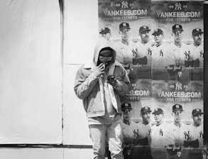A person wearing a hoodie and sunglasses stands in front of a wall plastered with multiple posters of a baseball team in uniform, promoting Yankees tickets for the 2023 season.