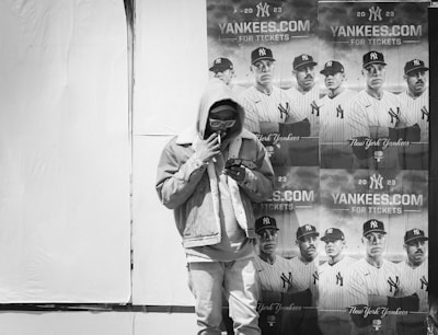 A person wearing a hoodie and sunglasses stands in front of a wall plastered with multiple posters of a baseball team in uniform, promoting Yankees tickets for the 2023 season.