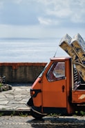 A small, orange three-wheeled vehicle is parked on a stone-paved area near a body of water. The vehicle is loaded with several large packages wrapped in blue and white paper. A brick and concrete barrier separates the vehicle from the water, and the sky is cloudy with patches of sunlight filtering through.
