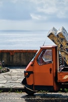 A small, orange three-wheeled vehicle is parked on a stone-paved area near a body of water. The vehicle is loaded with several large packages wrapped in blue and white paper. A brick and concrete barrier separates the vehicle from the water, and the sky is cloudy with patches of sunlight filtering through.