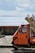 A small, orange three-wheeled vehicle is parked on a stone-paved area near a body of water. The vehicle is loaded with several large packages wrapped in blue and white paper. A brick and concrete barrier separates the vehicle from the water, and the sky is cloudy with patches of sunlight filtering through.