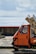 A small, orange three-wheeled vehicle is parked on a stone-paved area near a body of water. The vehicle is loaded with several large packages wrapped in blue and white paper. A brick and concrete barrier separates the vehicle from the water, and the sky is cloudy with patches of sunlight filtering through.