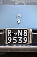 A close-up view of a vintage car's license plate with 'ROMA N8 9539' in white numbers and letters on a black background. Above the license plate, there is a car emblem shaped like two chevrons.