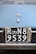 A close-up view of a vintage car's license plate with 'ROMA N8 9539' in white numbers and letters on a black background. Above the license plate, there is a car emblem shaped like two chevrons.