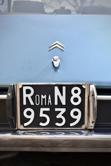 A close-up view of a vintage car's license plate with 'ROMA N8 9539' in white numbers and letters on a black background. Above the license plate, there is a car emblem shaped like two chevrons.