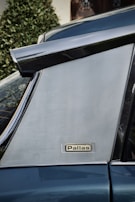 Close-up of a metallic car surface featuring a chrome trim and badge labeled 'Pallas'. The background includes a blurred view of a shrub and a window with a curtain.