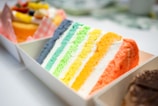 A slice of layered cake with bright, colorful stripes. The layers are arranged in a sequence of colors ranging from blue to green, yellow, and orange. It is placed in a white container with other desserts blurred in the background.