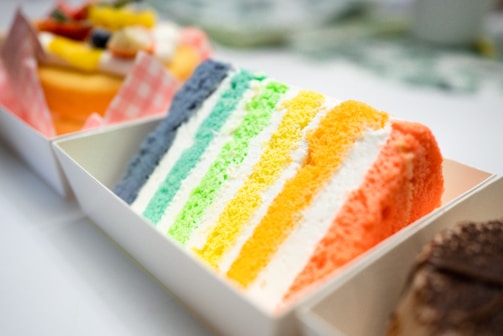 A slice of layered cake with bright, colorful stripes. The layers are arranged in a sequence of colors ranging from blue to green, yellow, and orange. It is placed in a white container with other desserts blurred in the background.