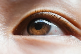 A close-up view of a human eye, focusing on the iris and eyelashes. The iris is brown with a reflective surface showing light patterns, while fine eyelashes frame the eye.