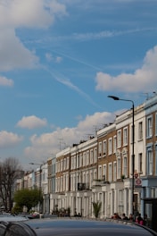 A row of classic terraced houses with white and brick facades extends along a street under a blue sky with scattered clouds. The street is lined with cars and pedestrians, featuring lampposts and trees that add greenery to the urban setting. Television antennas protrude from the rooftops, and a red circle sign is visible on the right.