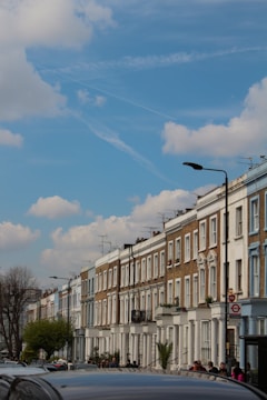 A row of classic terraced houses with white and brick facades extends along a street under a blue sky with scattered clouds. The street is lined with cars and pedestrians, featuring lampposts and trees that add greenery to the urban setting. Television antennas protrude from the rooftops, and a red circle sign is visible on the right.