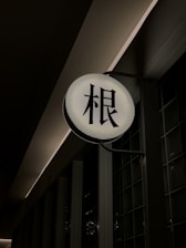 A round, illuminated sign with Japanese kanji characters in black against a bright white background. The sign is mounted on a dark wall, slightly protruding out. The surrounding area is dimly lit, creating a contrast between the light of the sign and the shadows of the environment.