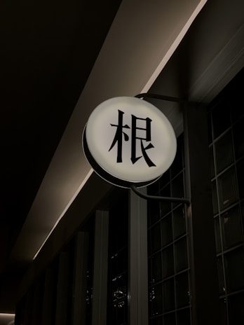 A round, illuminated sign with Japanese kanji characters in black against a bright white background. The sign is mounted on a dark wall, slightly protruding out. The surrounding area is dimly lit, creating a contrast between the light of the sign and the shadows of the environment.
