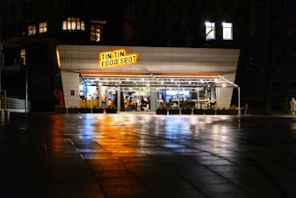 A modern, well-lit eatery named 'Tin Tin Food Spot' is located on a dimly lit street. The establishment features large glass windows through which the interior and seating arrangements are visible. The pavement reflects light, suggesting that it is slightly wet. A solitary person dressed in light clothing walks near the restaurant.