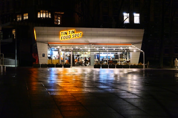 A modern, well-lit eatery named 'Tin Tin Food Spot' is located on a dimly lit street. The establishment features large glass windows through which the interior and seating arrangements are visible. The pavement reflects light, suggesting that it is slightly wet. A solitary person dressed in light clothing walks near the restaurant.