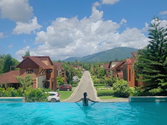 A scenic view featuring a person relaxing in an infinity pool, overlooking a row of brown-roofed houses, green foliage, and a distant mountainous landscape under a bright blue sky with scattered clouds.