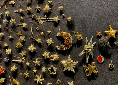 A collection of star-themed pins and brooches arranged on a dark textured surface. The items vary in design, featuring stars, crescent moons, and other celestial shapes, some adorned with gemstones or colorful enamel.