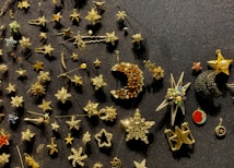 A collection of star-themed pins and brooches arranged on a dark textured surface. The items vary in design, featuring stars, crescent moons, and other celestial shapes, some adorned with gemstones or colorful enamel.