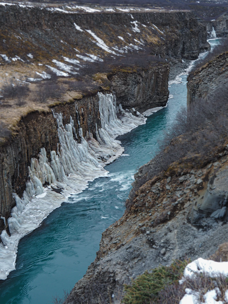 The image depicts a breathtaking view of a river flowing through a mountainous landscape. The river, with its deep blue waters, is surrounded by snow-capped mountains on both sides. The waterway is narrow and winding, adding to the sense of adventure in the scene. On one side of the river, there are trees and shrubs, while on the other side, rocks and boulders can be seen. The sky above is overcast, casting a soft light over the entire landscape.